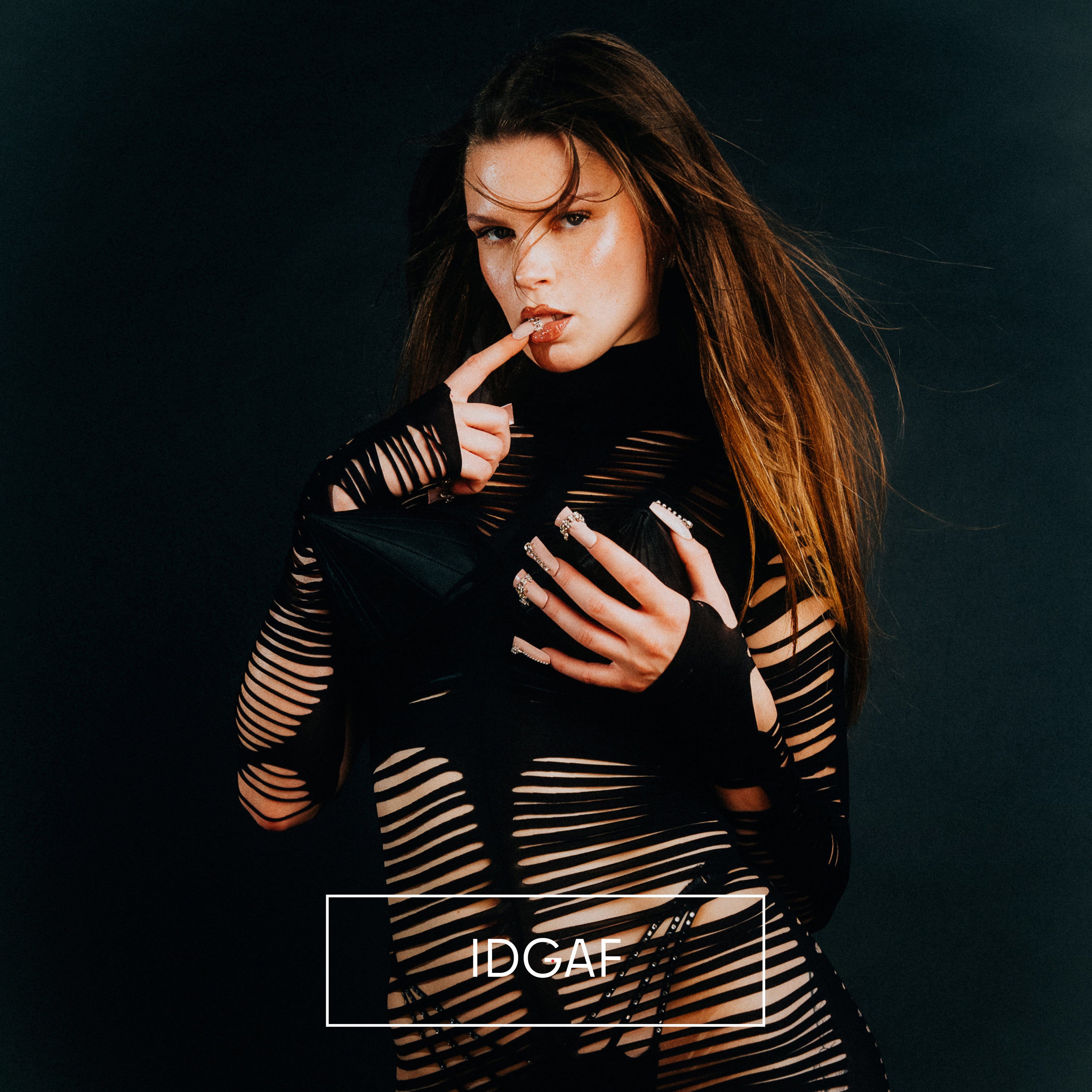 „Covermotiv mit einer Frau vor dunklem Hintergrund, die ein schwarzes, zerrissenes Outfit mit Cut-outs trägt. Sie blickt direkt in die Kamera und legt einen Finger an die Lippen. Unten steht der Schriftzug ‚IDGAF