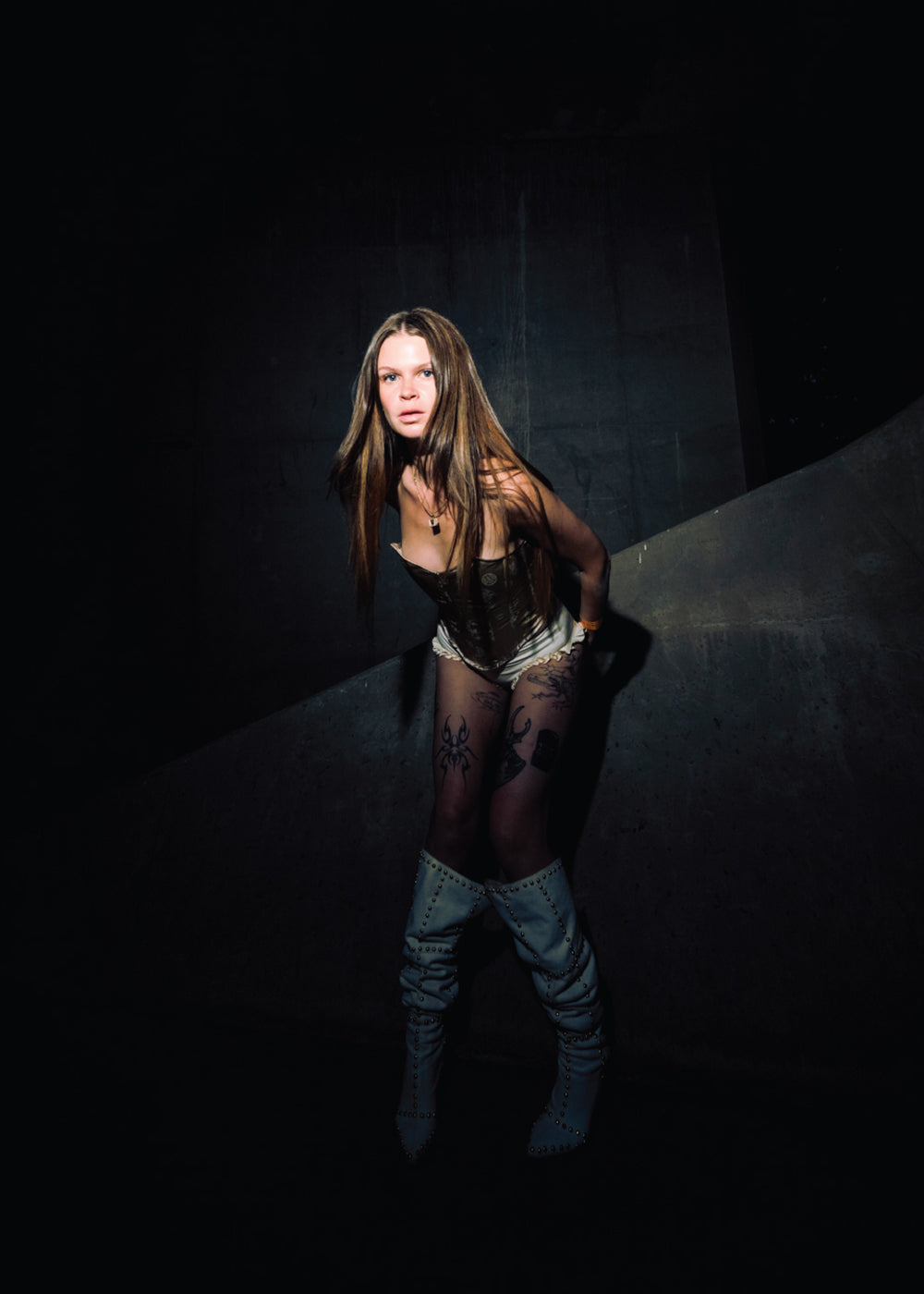„Person mit langen Haaren trägt ein Korsett, helle Shorts und hohe weiße Stiefel. Das Motiv ist in dunklem Licht aufgenommen, mit Betonwand im Hintergrund.“